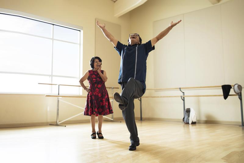 A Hispanic man leaps for joy in a dance studio while his wife smiles on.